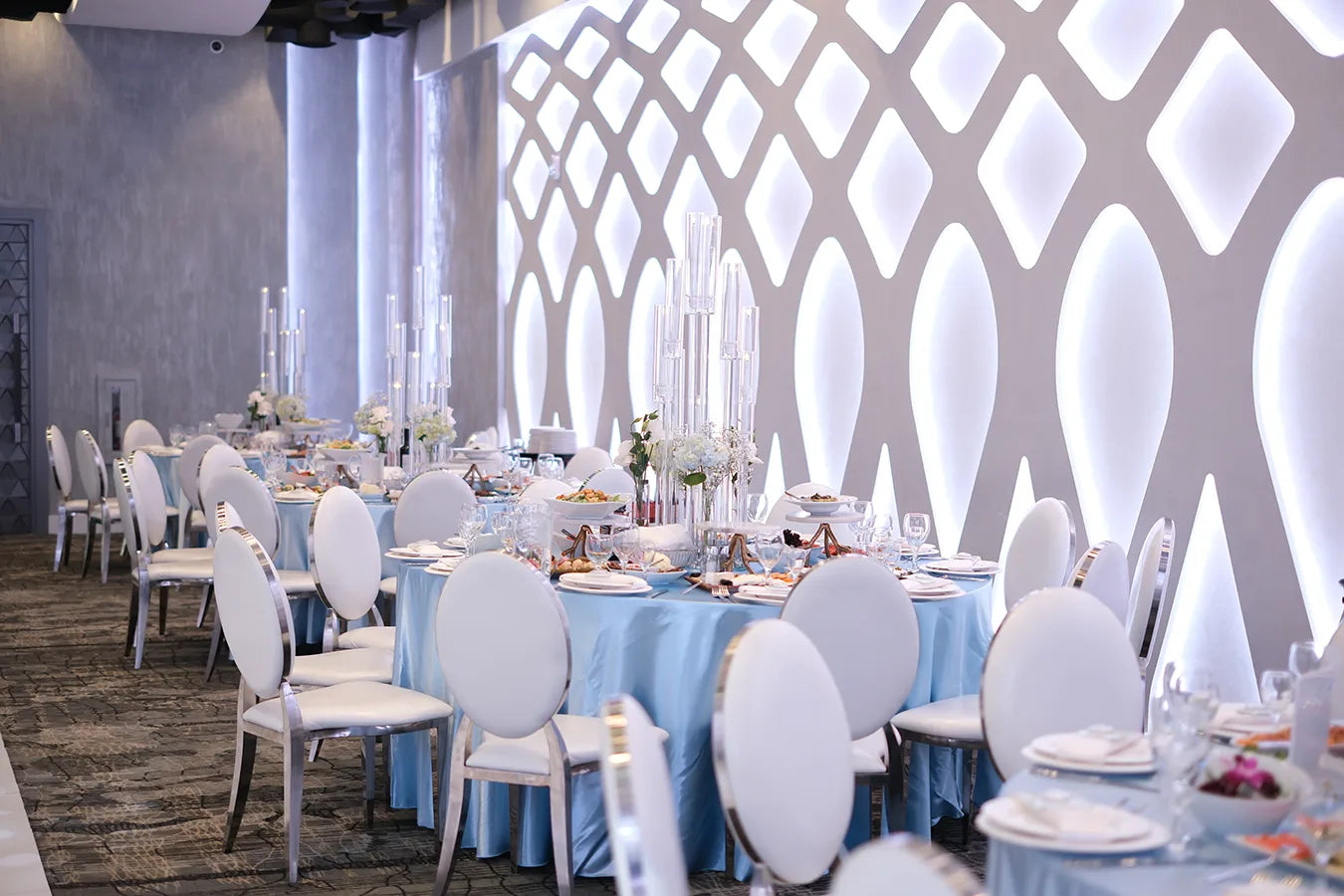 Elegant banquet hall with round tables covered in light blue tablecloths, white chairs, tall glass centerpieces, floral arrangements, and a modern, illuminated geometric wall in the background.