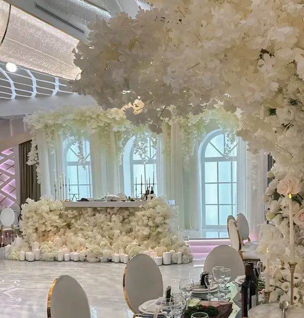 A lavishly decorated banquet hall features white floral arrangements, tall arched windows, elegant round chairs, and a table with candles, creating a luxurious and romantic atmosphere.