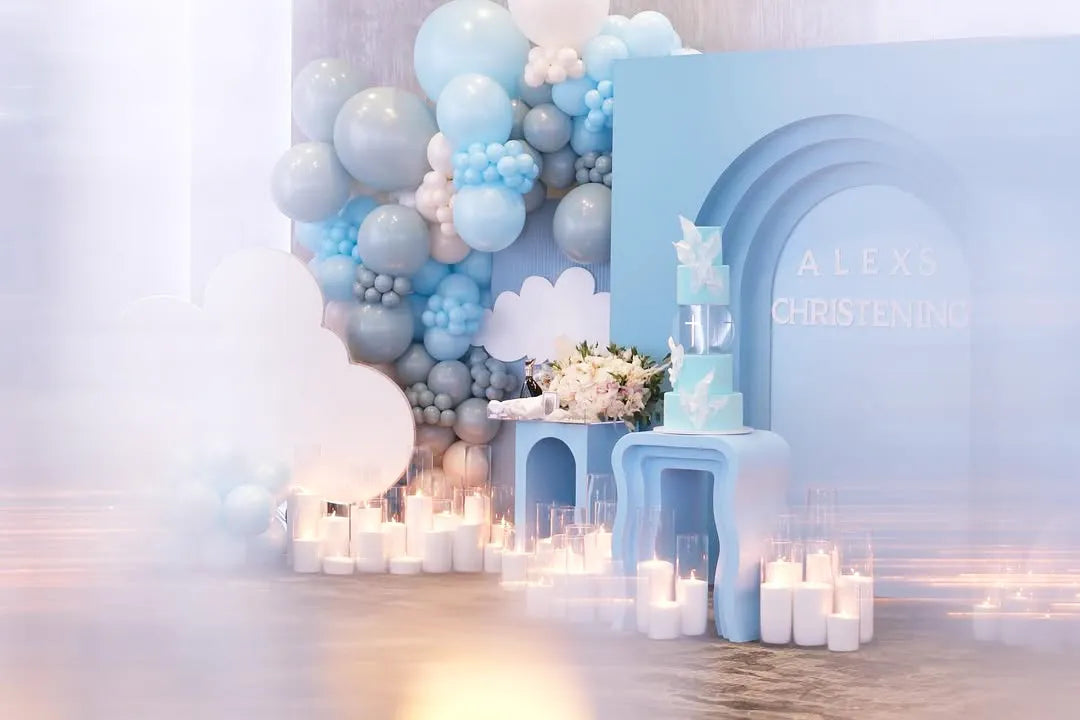 A decorative setup for a christening with blue and white balloons, candles, flowers, and a blue arch with “Alex’s Christening” written on it. A tiered cake and cloud-shaped props complete the scene.