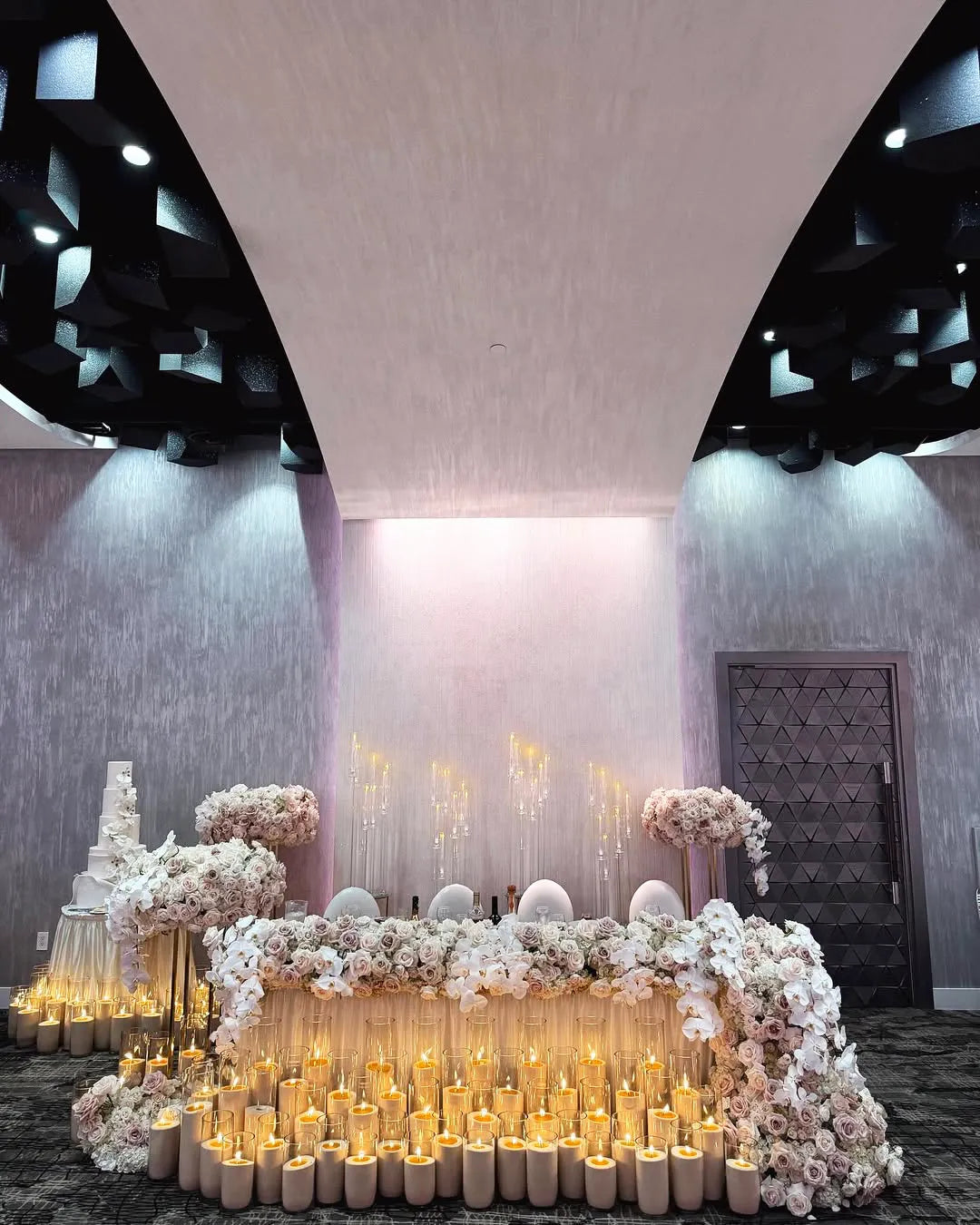 A lavish wedding reception table decorated with white and blush flowers, dozens of lit candles in glass holders, and a tall white cake. The setting features modern gray walls, a black geometric ceiling, and elegant lighting.
