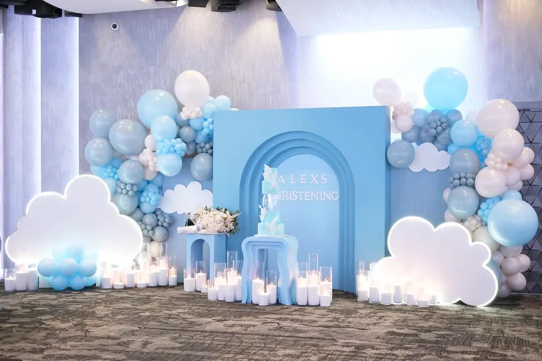 A pastel blue and white christening party setup features balloon garlands, cloud-shaped cutouts, and many candles. A blue backdrop with 