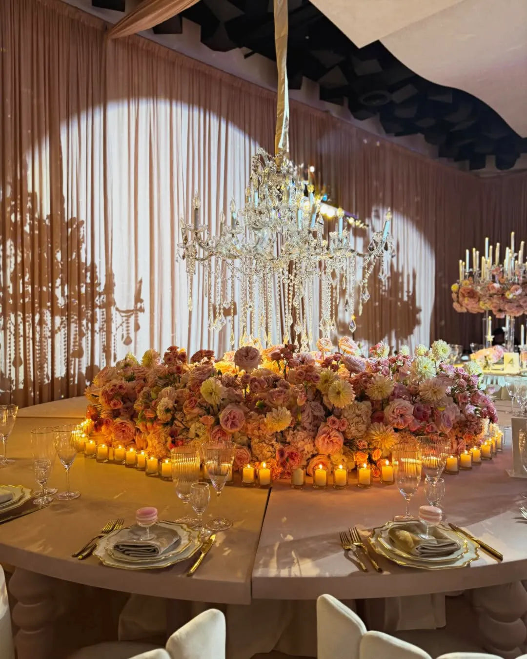 A lavish banquet table is set with gold flatware, crystal glassware, and gold-rimmed plates. The centerpiece is a large arrangement of pink and cream flowers, surrounded by candles, beneath a sparkling crystal chandelier.