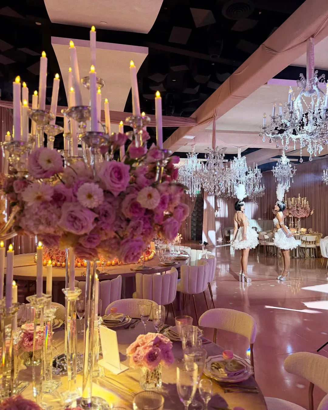 Elegant ballroom decorated with pink and purple floral centerpieces, crystal chandeliers, and candlelight. Two ballerinas in white tutus stand in the background, adding a whimsical touch to the luxurious setting.