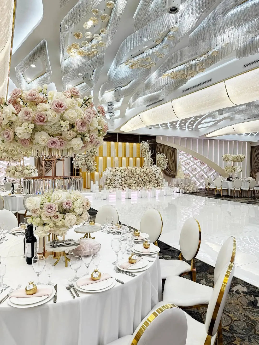 Elegant wedding reception hall with round tables set for guests, decorated with large floral centerpieces of white and blush roses. The room features a glossy white floor and a modern ceiling with golden and white accents.