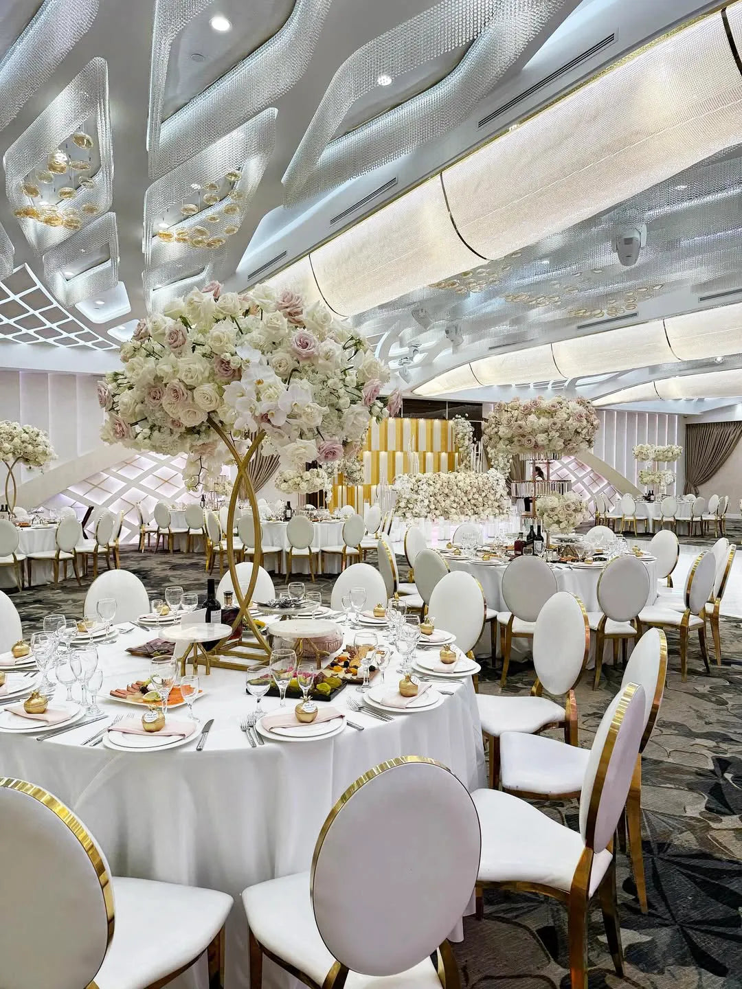 Elegant banquet hall with round tables set for an event, featuring white tablecloths, gold-accented chairs, and tall floral centerpieces with white and blush flowers under modern, decorative ceilings.