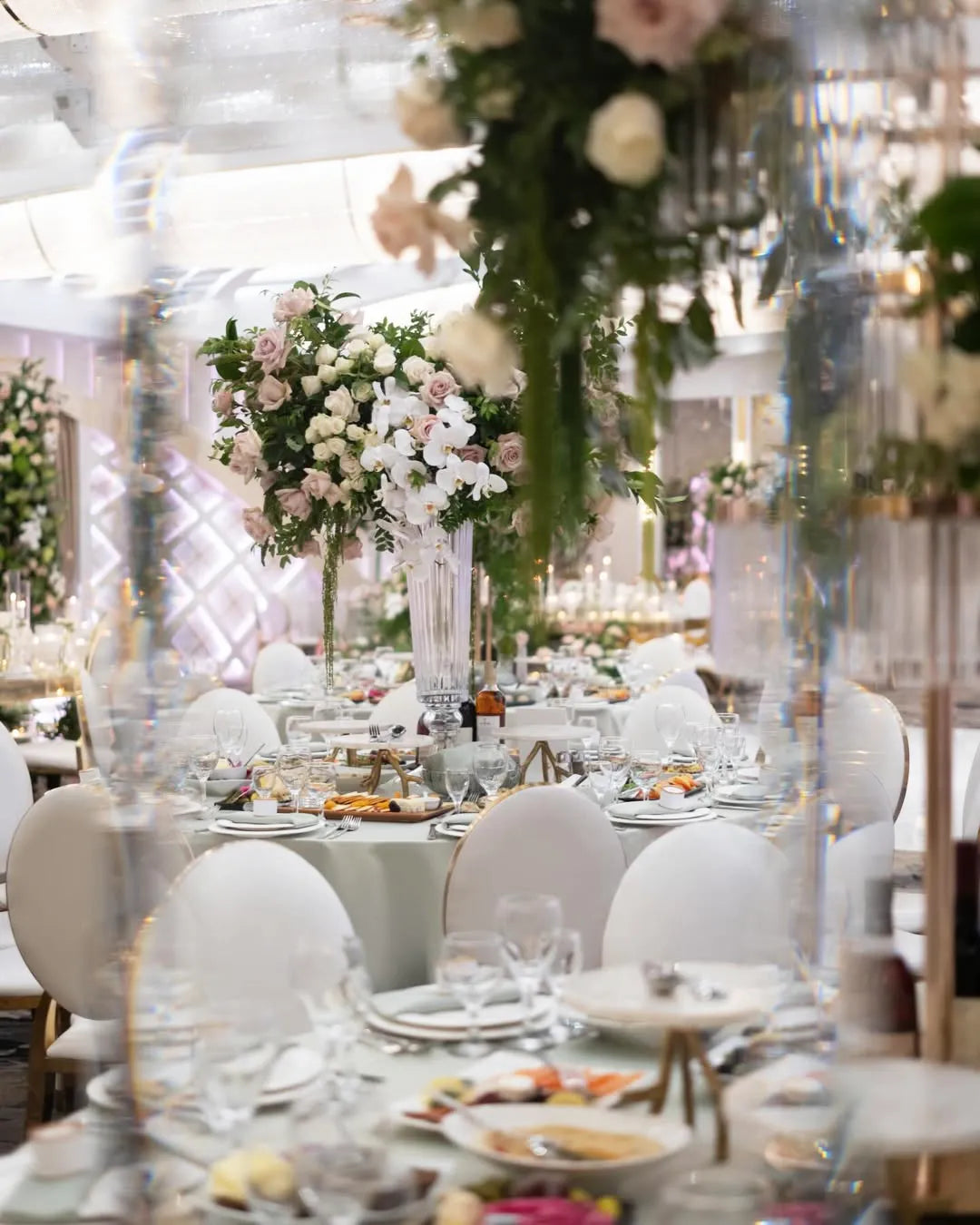 Elegant banquet hall with round tables set for an event, featuring white chairs, neatly arranged plates, glasses, and cutlery. Tall floral centerpieces with white and pink flowers add a luxurious touch to the sophisticated setting.