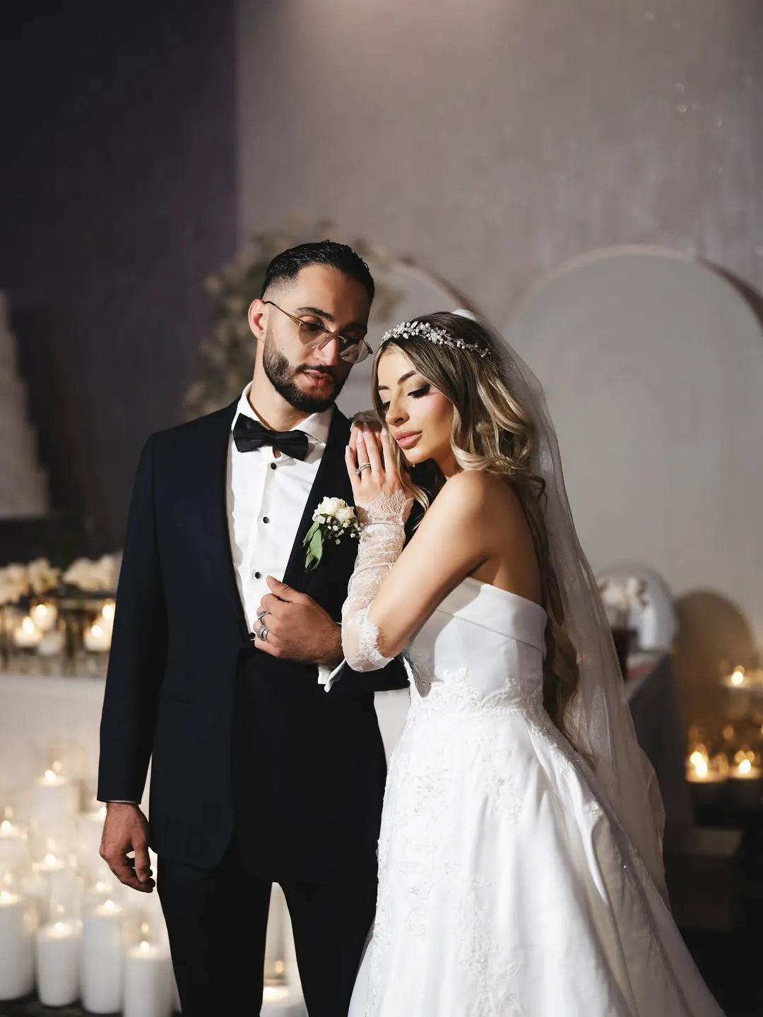A bride in a white gown and veil leans gently on the shoulder of a groom in a black tuxedo, surrounded by glowing candles in a softly lit, elegant setting.
