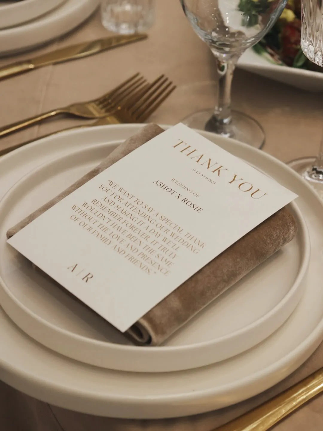 A wedding thank you card rests on a folded taupe napkin atop a white plate, surrounded by gold cutlery and glassware on an elegantly set table.