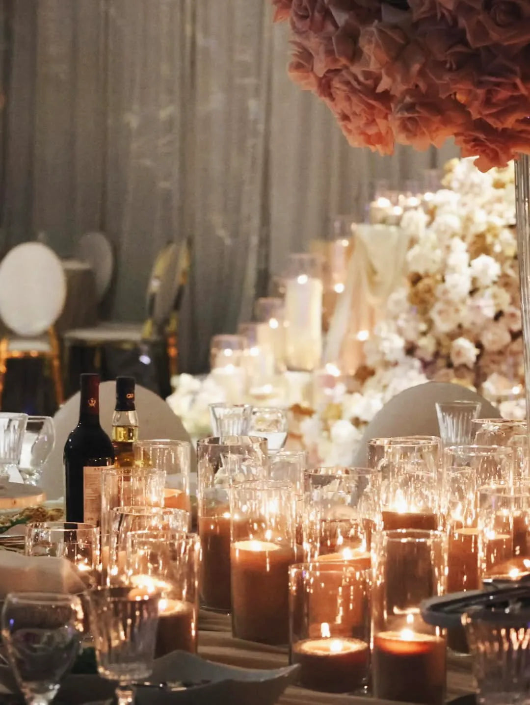 A beautifully decorated event table with numerous lit candles, wine bottles, glassware, and floral arrangements; soft lighting and a backdrop of stairs adorned with flowers and candles create an elegant, romantic atmosphere.