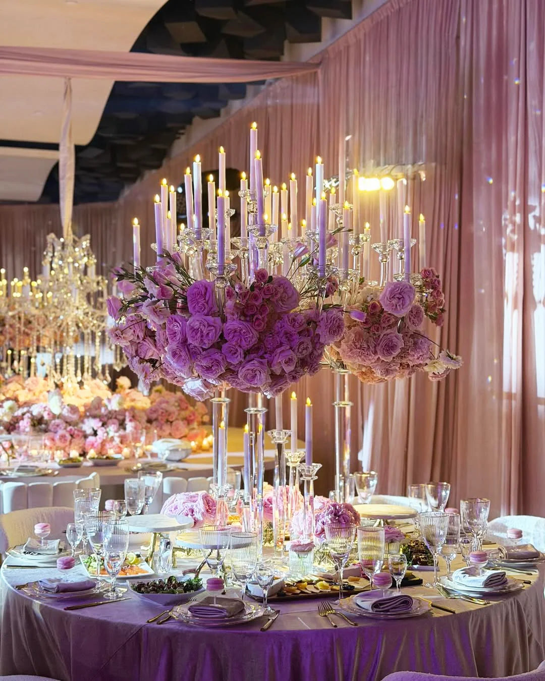 Elegant banquet table with a tall candelabra centerpiece adorned with pink and purple roses, surrounded by glassware, plates, and pink napkins. The room is decorated with pink drapes and soft lighting, creating a luxurious ambiance.