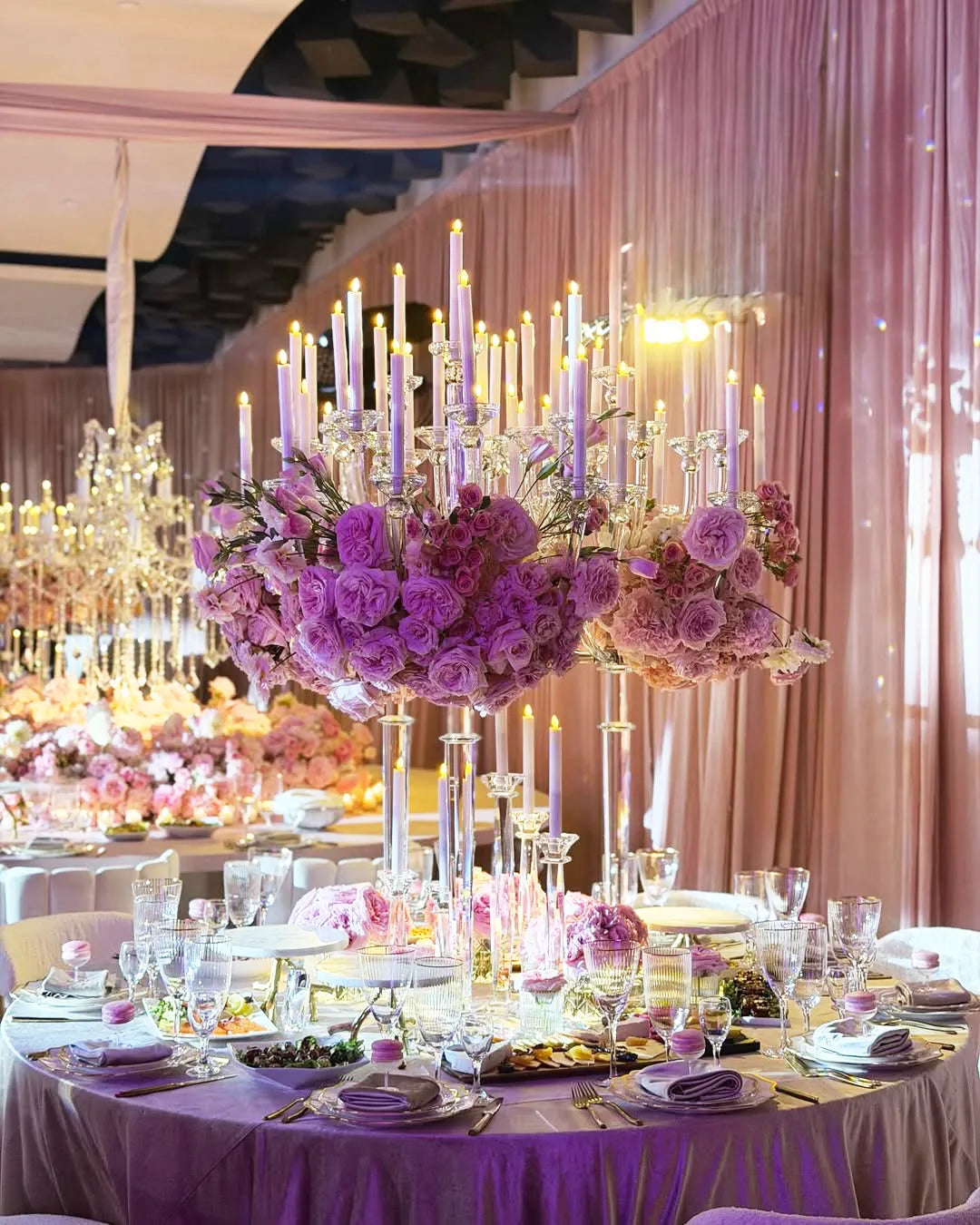 A lavishly decorated banquet table features tall crystal candelabras with lit candles, large arrangements of pink and purple flowers, elegant tableware, and plush pink drapes in the background, creating an opulent ambiance.