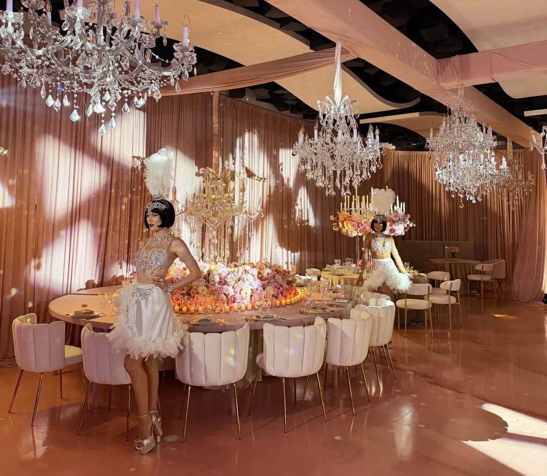 Opulent banquet hall with pink drapes, large chandeliers, flower arrangements, decorated round tables, and two women in ornate white feathered dresses standing near the tables.