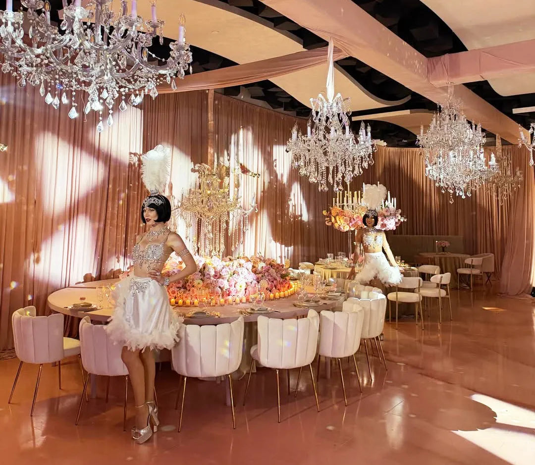 Two women in elegant, feathered white dresses stand in a luxurious banquet hall with chandeliers, pink drapes, floral centerpieces, and white chairs around a curved dining table. The room is bathed in soft pink lighting.