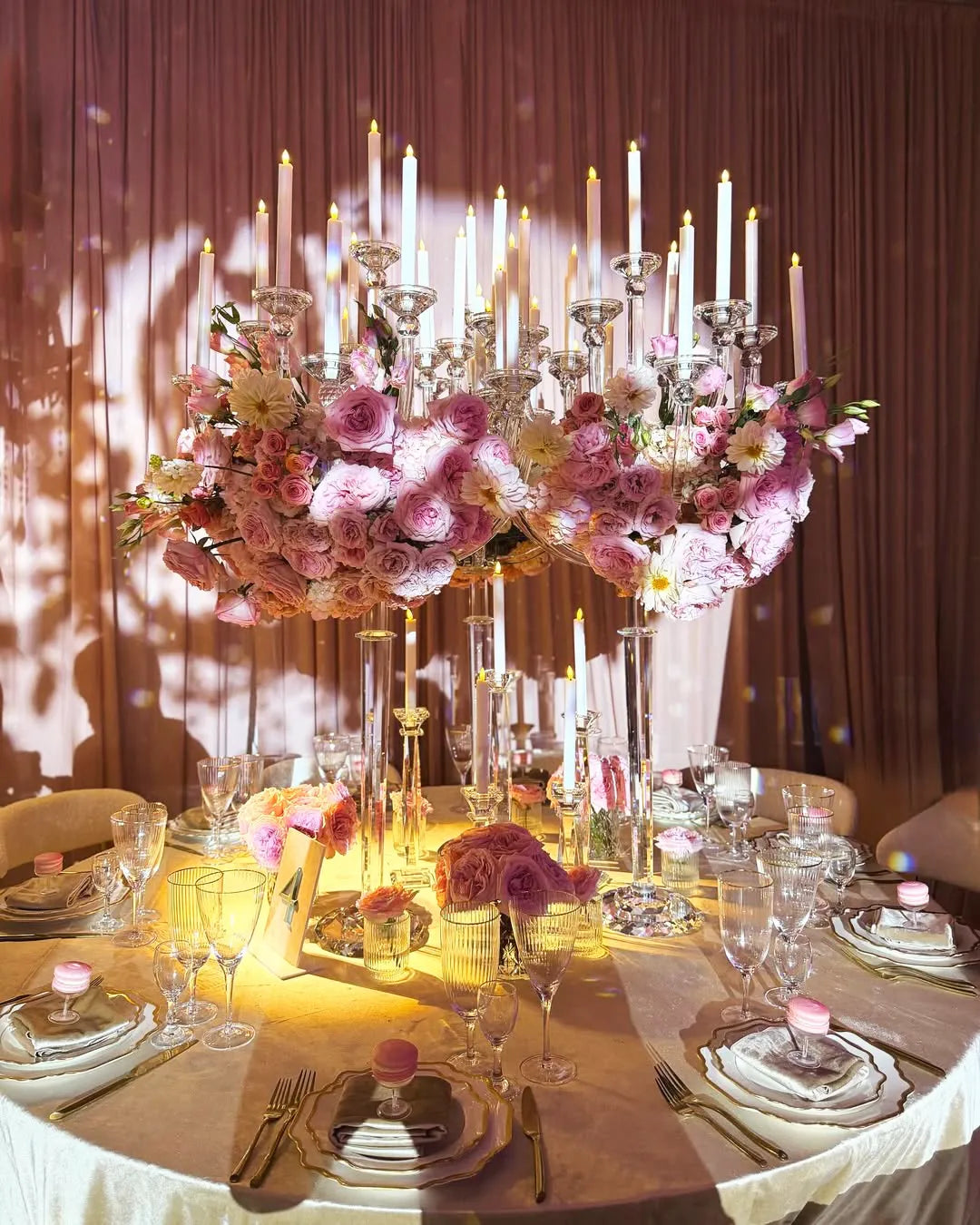 An elegant table setting features a large floral centerpiece with pink roses, tall white candles in glass holders, and fine china with pink macarons at each place, all bathed in warm, romantic lighting.