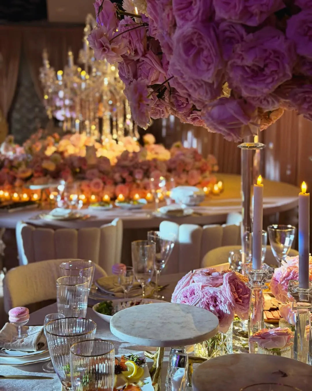Elegant banquet table set with glassware, plates, and floral centerpieces of pink roses; candles and chandeliers create a warm, romantic atmosphere, with more flower arrangements in the background.