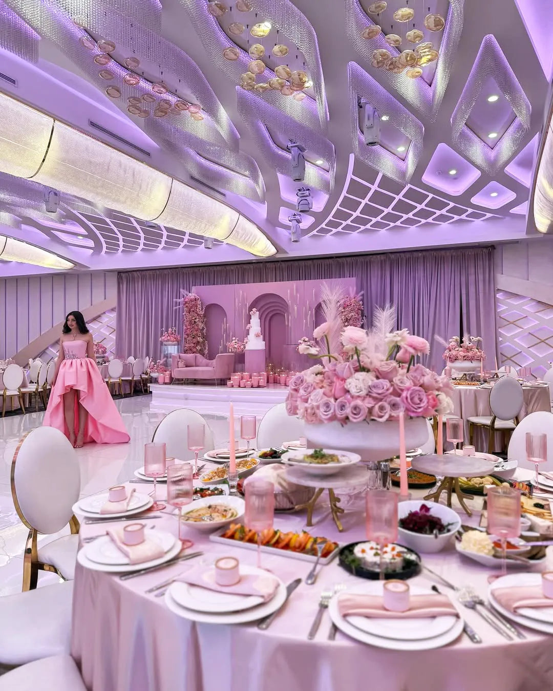 A lavish banquet hall decorated in pink and purple, featuring elegant round tables set with flowers, candles, and fine dining ware. The ceiling is ornate and illuminated. A woman in a pink dress stands in the background near a large cake display.