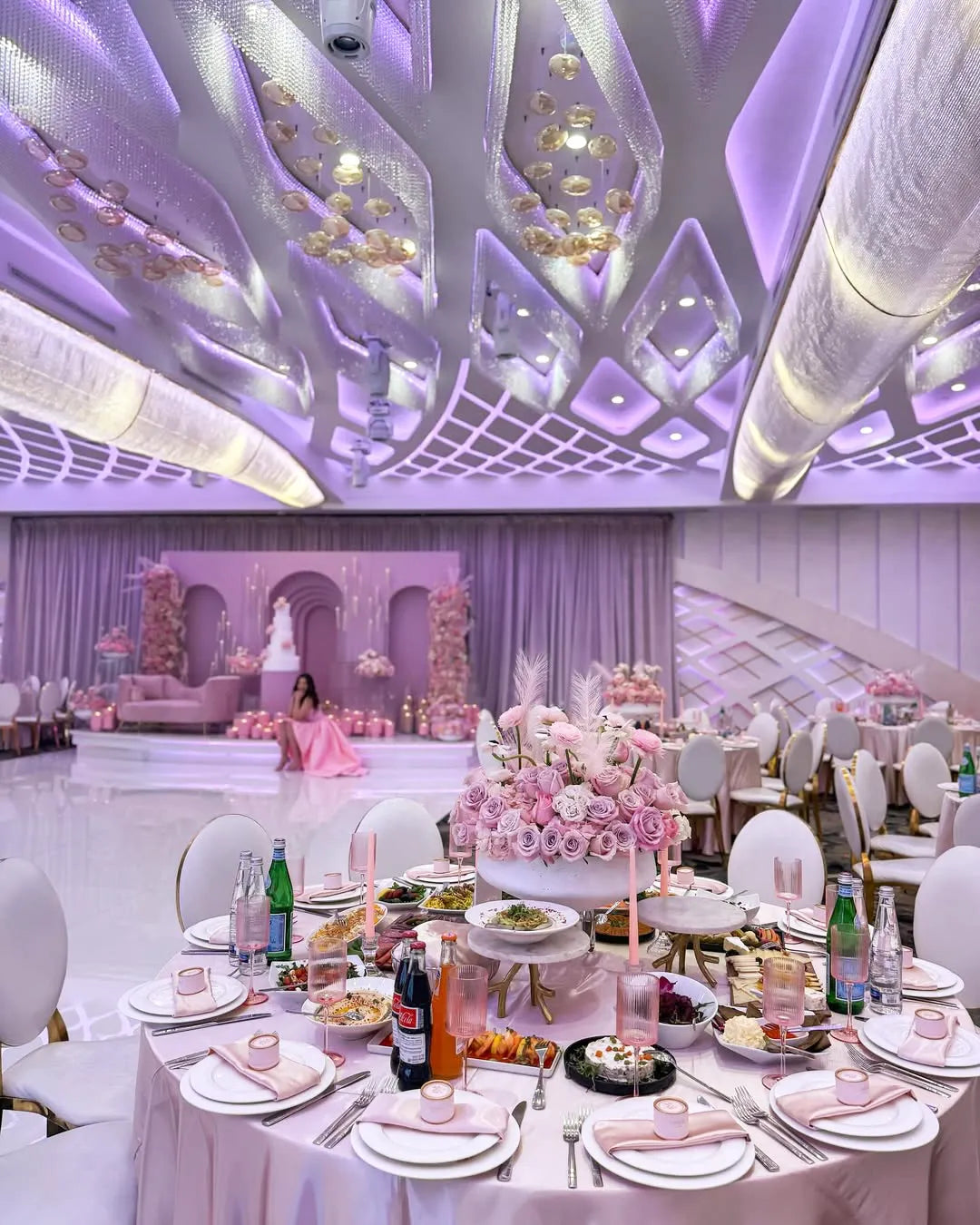 A lavish banquet hall decorated in pink and purple tones. Round tables with floral centerpieces, food, and drinks are set up. In the background, a woman in a pink dress sits on a stage adorned with flowers.
