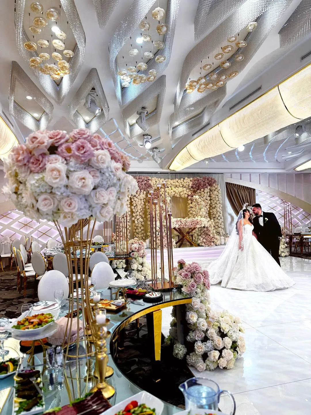 A bride and groom embrace on a luxurious, flower-filled wedding stage in an elegant, modern ballroom with elaborate ceiling decor and lavishly set tables adorned with pink and white floral arrangements.