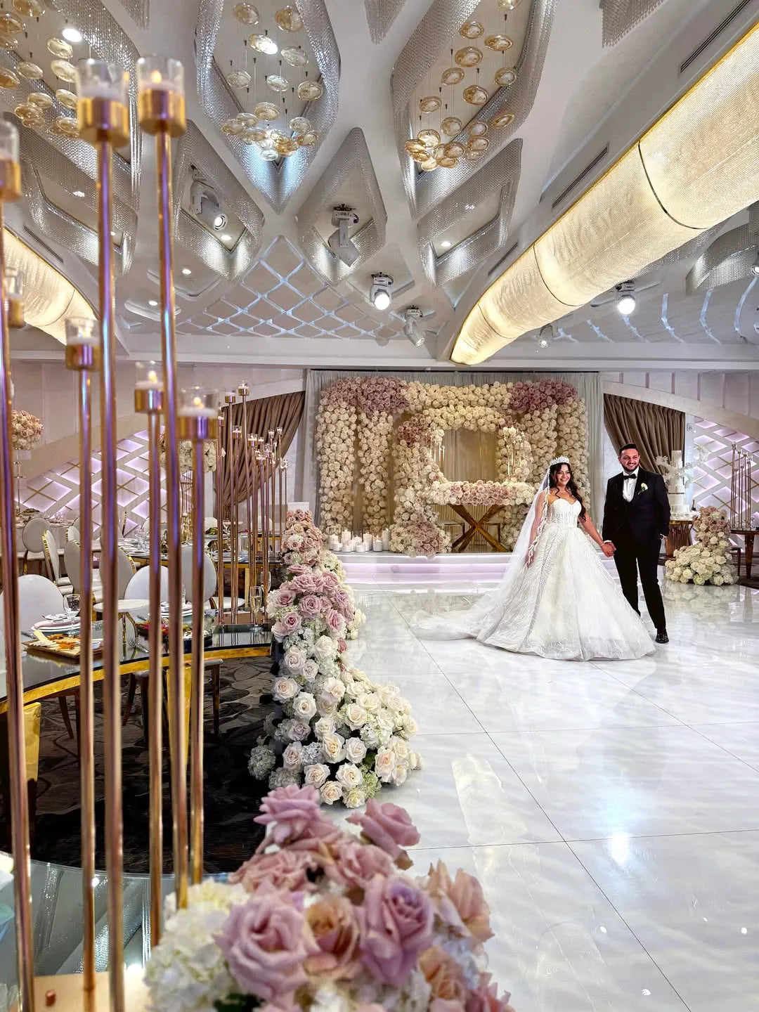 A bride and groom walk hand in hand in an ornate wedding hall decorated with white and blush flowers, gold accents, and elegant lighting. The ceiling has geometric designs, and a floral arch is in the background.