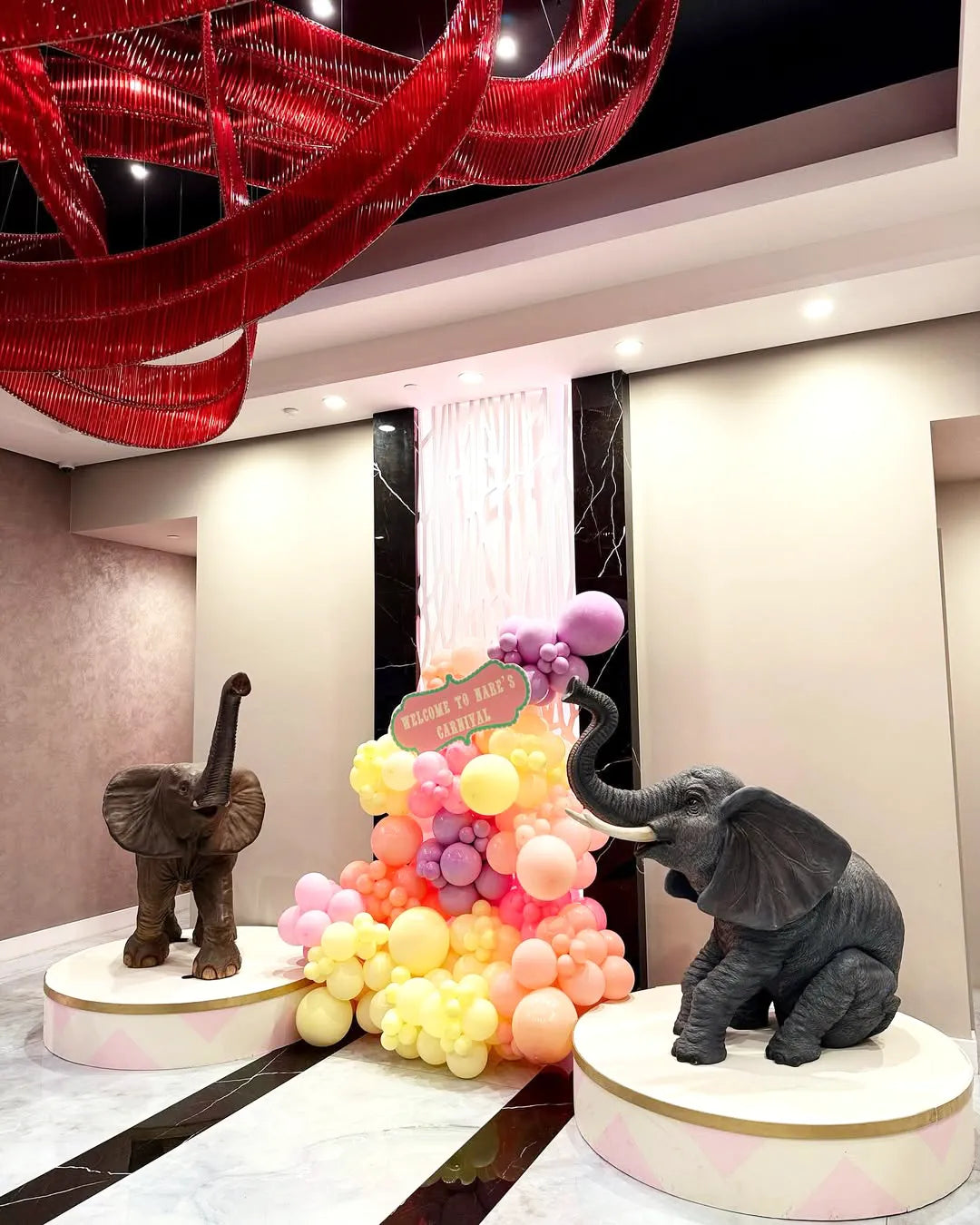 Two realistic elephant statues stand on round podiums, flanking a pastel balloon arrangement with a sign that reads, 