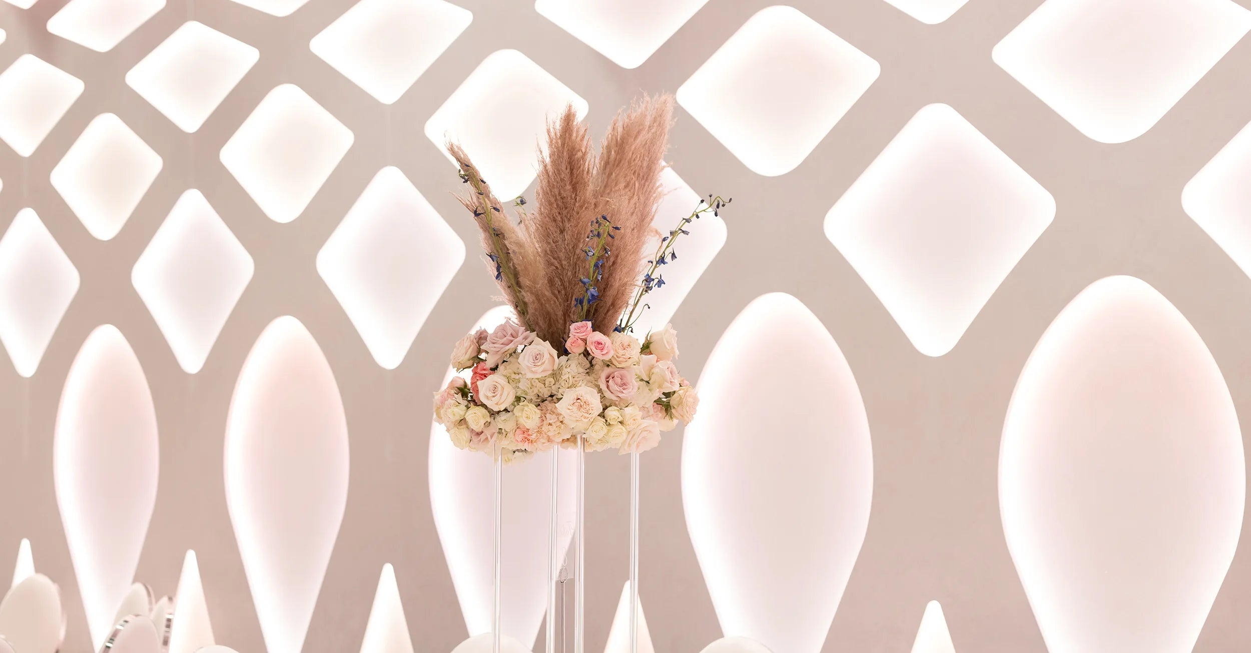 A modern floral arrangement with pampas grass, pink and white roses, and white flowers sits in front of a white geometric wall with glowing diamond-shaped patterns.