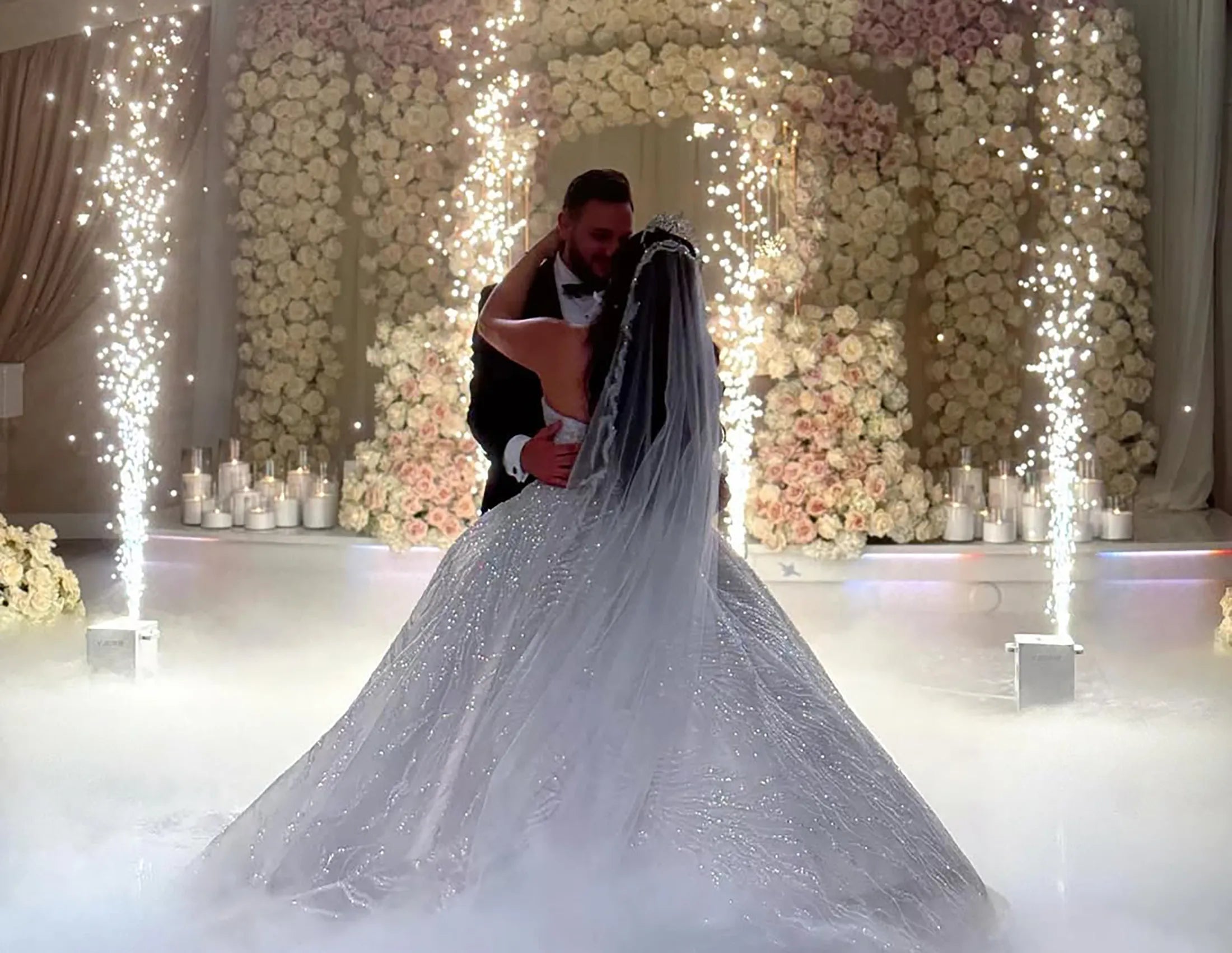 A bride and groom share their first dance surrounded by fog and sparkling fountains, with a floral arch and candles in the background, creating a magical wedding scene.