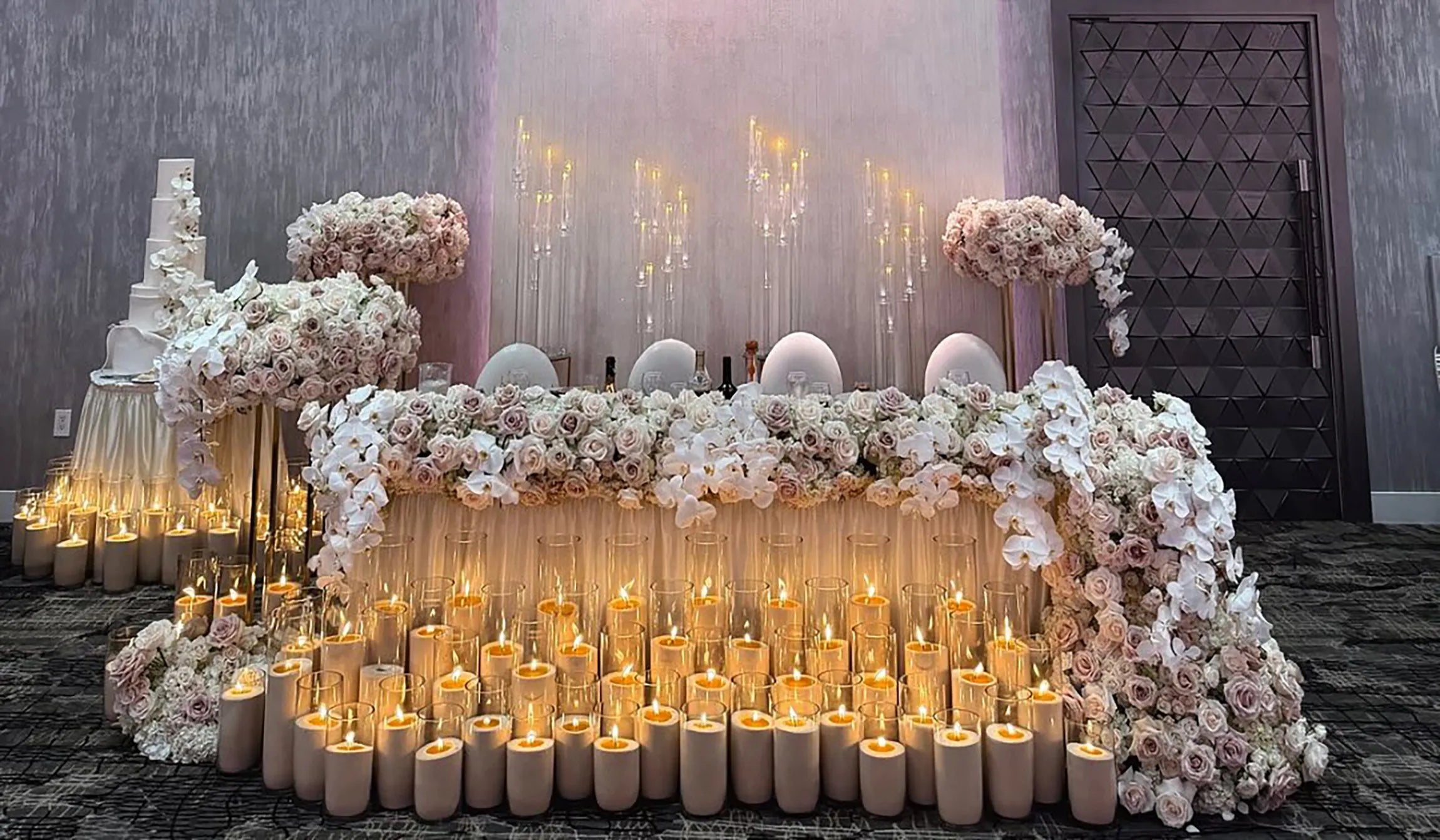 A wedding table decorated with white and pink flowers, surrounded by many lit candles in glass holders. The backdrop features draped fabric, soft lighting, and two large floral arrangements on either side.