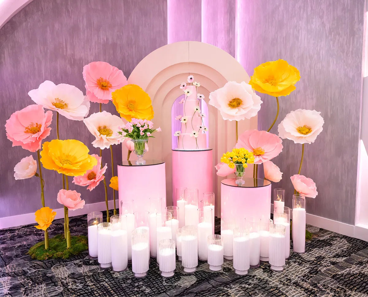 A decorative display featuring large pink and yellow paper flowers, three pink cylindrical stands with flowers, and many lit white candles on the floor, set against a light gray and pink background.