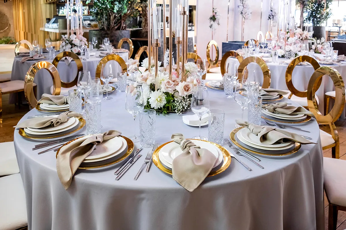 A round banquet table set for an event with gold-rimmed plates, beige napkins, clear glasses, gold chairs, and a floral centerpiece featuring white and pale pink flowers. The setting is elegant and formal.