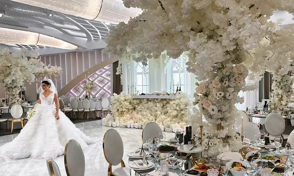 A bride in a white gown stands in an elegant, modern wedding venue decorated with large white floral arrangements, round tables set for guests, and silver chairs, creating a luxurious atmosphere.
